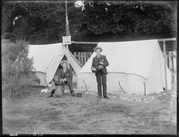 Image: Two unidentified young men in straw hats having breakfast, one pouring tea, one cooking over a wood burner, in front of two tents with an 'The Imperial Camp' sign and flag, trees beyond, [Sumner?], Christchurch