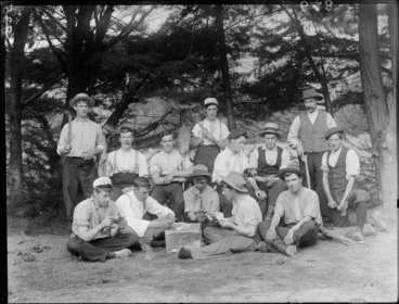 Image: A group portrait of the summer campers, showing some playing cards, one holding a tomahawk, one holding an axe, one holding a mallet, two holding brooms and one pouring beer into a glass and two smoking, probably Christchurch district