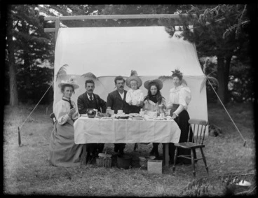 Image: Six unidentified people sitting at a table having a cup of tea, cakes and sandwiches in front of the tent, with a pot, a tin and two baskets with food in them under the table, showing two women, two men, a girl and a boy, possibly Christchurch district