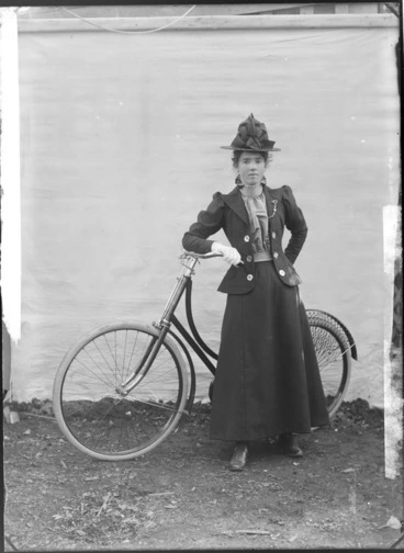 Image: Unidentified young woman, with a bicycle, outdoors, with a backdrop, probably Christchurch district