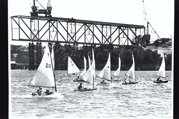Image: Group of children in P-class yachts sailing in front of construction work on the Auckland Harbour Bridge, 1950s