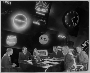Image: Showing six men working around a table with neon signs in the background, for Rayneon, 1941