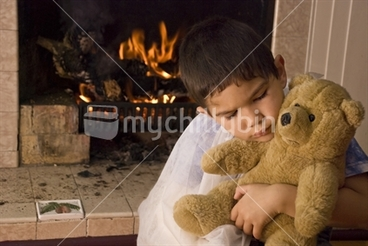 Image: A boy with a broken arm cuddles a teddy in front of a fire