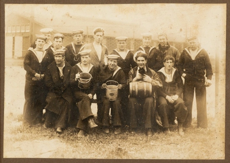 Image: Group of 14 sailors, two hold buckets and a third has a wooden cask on his knees and holds a cat in his hands. The ship name on the sailors' hats is hard to read but is possibly the HMS 'Chatham'.