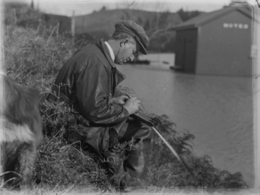 Image: [A man sitting on a bank beside a flooded area writing something]