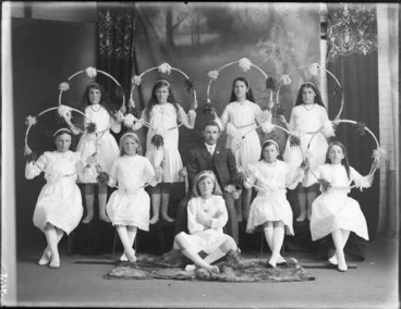 Image: Studio portrait of unidentified young girls in white dresses holding cane hula-loops decorated with small pompoms, arranged around a man [coach?], Christchurch