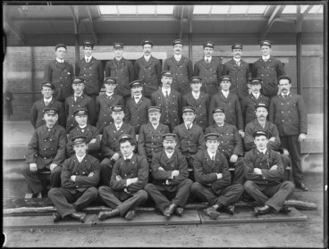 Image: Railway station unidentified workers group portrait, in uniform wearing caps with job function badges like porter or guard, under a railway station platform awning, Christchurch [Station?]