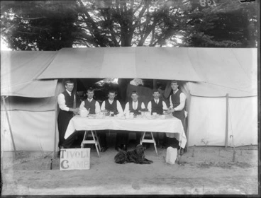 Image: Group of unidentified young men outside a tent having a tea party, probably Christchurch, includes a sign reading 'Tivoli Camp' and a dog resting in front