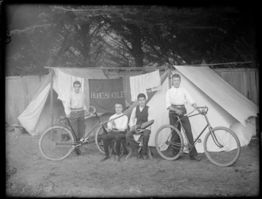 Image: Four unidentified boys, two with bicycles and two with flute and autoharp, in front of two tents with washing line between with 'Honeysuckle' banner, wooden fence and trees beyond, [Sumner?], Christchurch