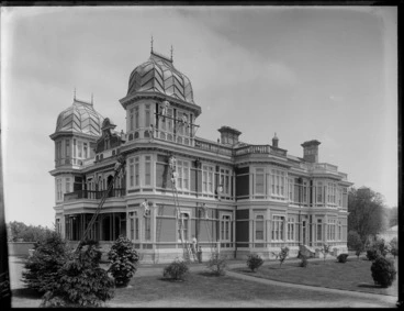 Image: McLean's Mansion, an elegant two story wooden building with twin ogival domes, showing painters with ladders and scaffolding, with well kept lawn and shrubs in front, Christchurch