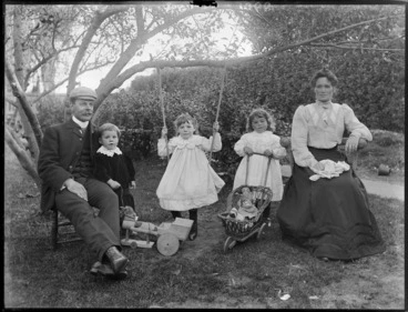 Image: Unidentified family group outdoors, showing couple and three children, one girl on swing, another girl with doll in pram and boy has a toy horse and cart, probably Christchurch district