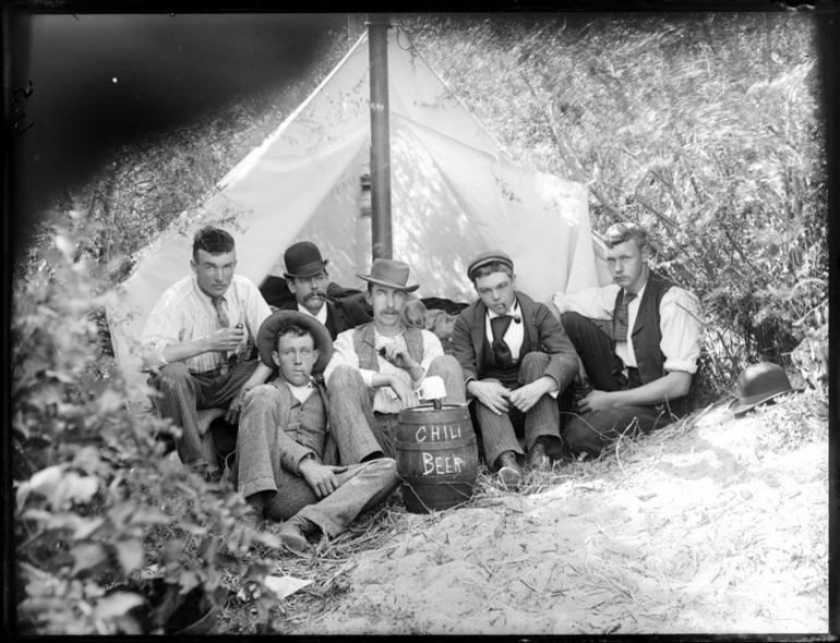 Image: Group of unidentified male youths outside tent, all with pipes, and a barrel which has the words 'Chili Beer' written on it with cup on top, probably Christchurch district