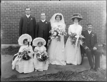Image: Wedding party portrait on long grass in front of brick wall, unidentified bride with long veil and groom, bridesmaid with large hat, best men and flower girls with large bonnets, probably Christchurch region