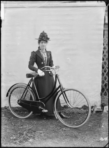 Image: Unidentified young woman, with a bicycle, outdoors, with a backdrop, probably Christchurch district