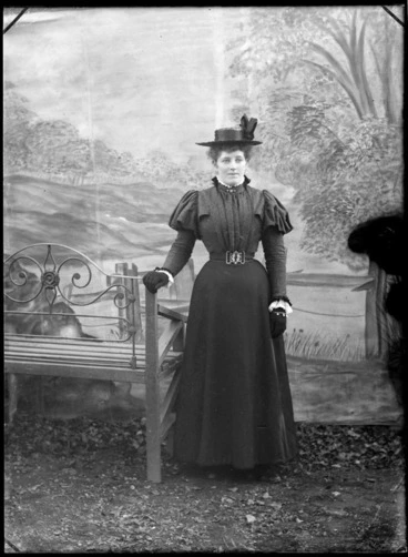 Image: Studio portrait of an unidentified woman standing next to a bench with painted backdrop, probably Christchurch district