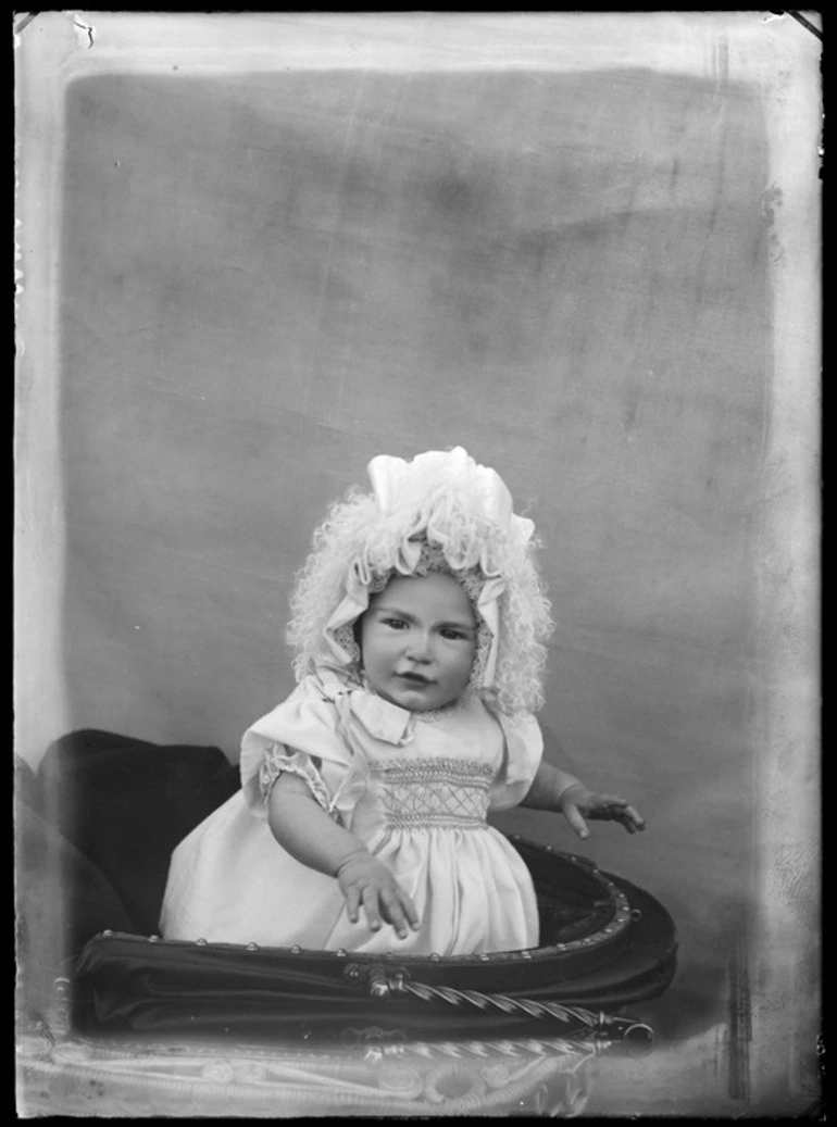 Image: Unidentified baby in a pram wearing a smocked dress and bonnet which has lace, ribbon and [hair extentions?] attached to it, probably Christchurch district Christchurch district