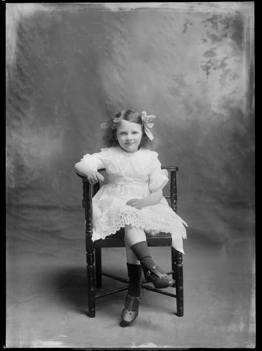 Image: Studio unidentified family portrait of a young girl in a lace dress with a large collar, belt and hair bow, sitting on a wooden chair, Christchurch