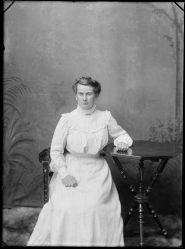 Image: Studio portrait of unidentified young woman in frilly lace white dress with high neck collar, sitting on a wooden chair next to a table, Christchurch