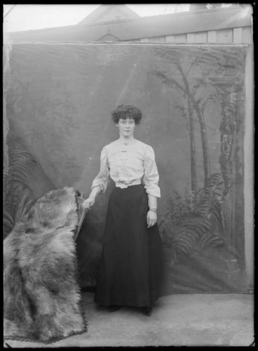 Image: Outdoors portrait in front of false backdrop, an unidentified young woman wearing high neck pleated shirt and sleeves, cameo brooch and chain bracelet, probably Christchurch region