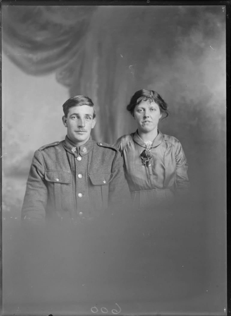 Image: Studio upper torso portrait of unidentified young WWI soldier with collar badges and black cord on left shoulder, standing with a young woman with brooch [sister, wife?], Christchurch