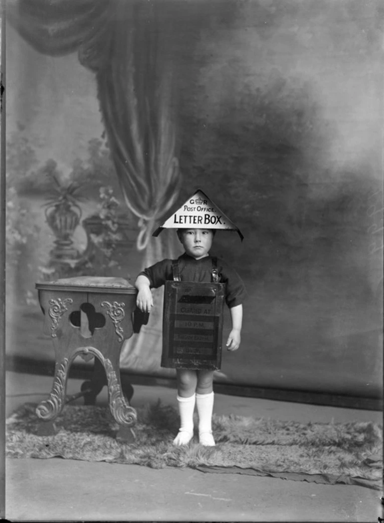 Image: Studio portrait of an unidentified boy in fancy dress as a GR Post Office Letterbox, showing the boy standing next to a wooden high chair and resting his right arm on it, possibly Christchurch district