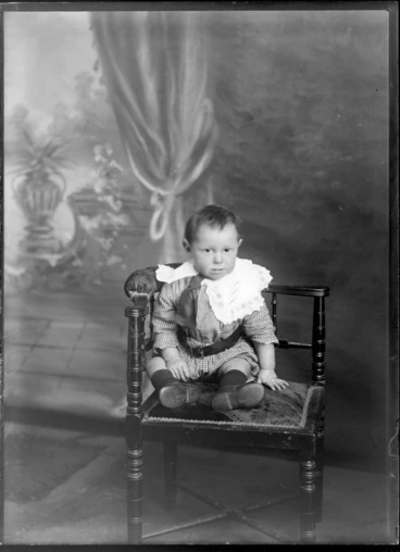 Image: Studio portrait of an unidentified boy dressed in a striped romper suit, with a large lace collar, tie and large belt, sitting on a wooden chair, possibly Christchurch district