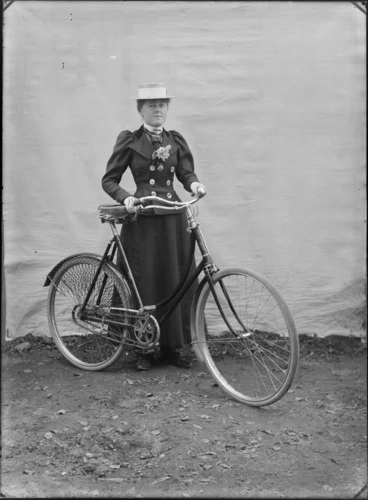 Image: Outdoors portrait with false backdrop, an unidentified woman in dark double breasted jacket with large white buttons and flowers, tie and straw hat, standing holding women's bicycle, probably Christchurch region