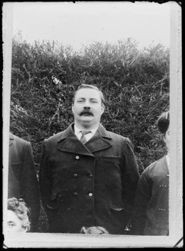 Image: Outdoors upper torso portrait of unidentified older man with large moustache and double breasted overcoat in front of tall hedge, people surrounding, probably Christchurch region
