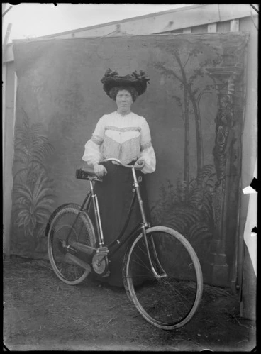 Image: Outdoors portrait in front of false backdrop, unidentified woman with lace blouse, necklace and large hat with ribbons, standing with women's bicycle, probably Christchurch region