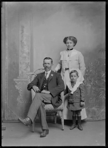 Image: Studio unidentified family portrait, father with moustache sitting, mother with portrait brooch and neck ribbon standing with young boy with large crocheted shoulder shawl, Christchurch