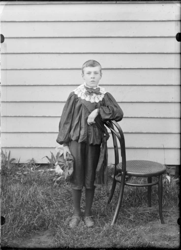 Image: Outdoor portrait of an unidentified boy dressed in knickerbockers, shirt with a lace collar, bow tie and sash, holding a hat in his left hand and leaning against a wooden chair, standing in front of a wooden building, possibly Christchurch district