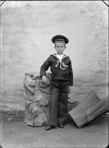 Image: Outdoor portrait of an unidentified boy dressed in a sailor outfit, standing in front of a studio canvas backdrop between a sack covered box and a tree stump, showing the words HMS Nautilus on his cap, possibly Christchurch district