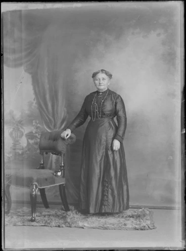Image: Studio portrait, unidentified elderly woman in dark dress with lace collar and buttons, neck chain, bar brooch with stones and a portrait brooch of a man, standing next to wooden padded chair, Christchurch