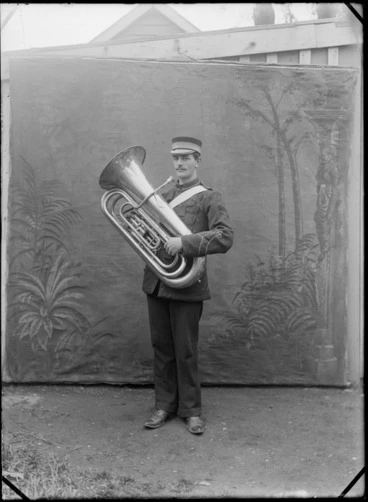 Image: Outdoors portrait in front of false backdrop, unidentified brass band player in uniform with sleeve insignia and hat, with handlebar moustache and tuba, probably Christchurch region