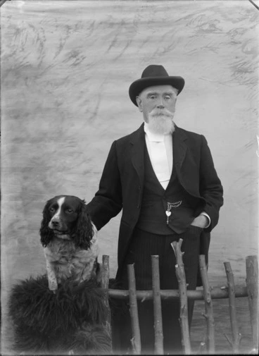 Image: Outdoors portrait in front of false backdrop, unidentified elderly man with goatee beard and hat with wide white tie, standing with Cocker Spaniel dog, probably Christchurch region