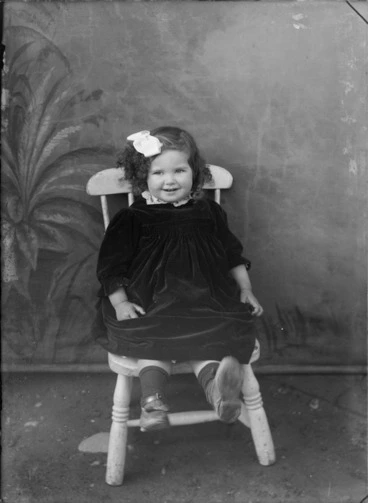 Image: Outdoors family portrait in front of false backdrop, an unidentified young girl in dark felt dress and white lace collar, sitting on small wooden chair, Christchurch