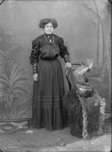Image: Outdoors portrait in front of false backdrop, an unidentified woman in high neck dark lace embroidered dress, standing with chair and potted plant, probably Christchurch region