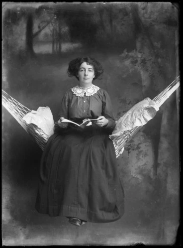 Image: Studio portrait of unidentified woman, sitting on a hammock with cushions and reading a book, probably Christchurch district