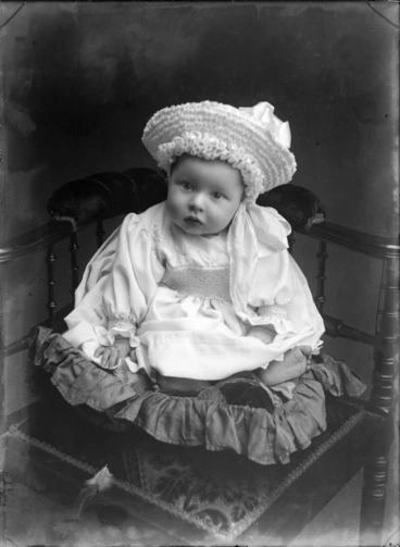 Image: Studio portrait of unidentified baby, wearing a bonnet and sitting on a cushion, probably Christchurch district