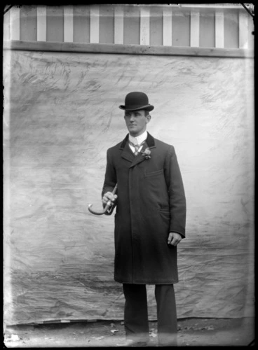 Image: Unidentified man wearing a bowler hat and holding a cane outdoors, with a painted studio backdrop, probably Christchurch district