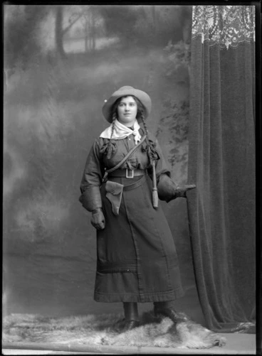 Image: Studio portrait of an unidentified young woman, wearing a 'Wild West' costume, including leather gauntlets, gun holster, with a handkerchief tied around neck, a whip across chest, and plaited pigtails, possibly Christchurch district