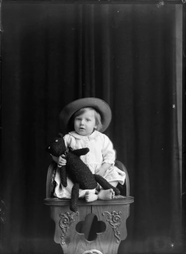 Image: Studio family portrait of unidentified young girl with large hat and holding a teddy bear while sitting on wooden high chair, Christchurch