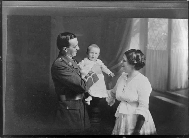 Image: Family unidentified portrait, front room of house with bay windows, a World War soldier with collar badges and sleeve insignia holding baby with wife looking on, probably Christchurch region