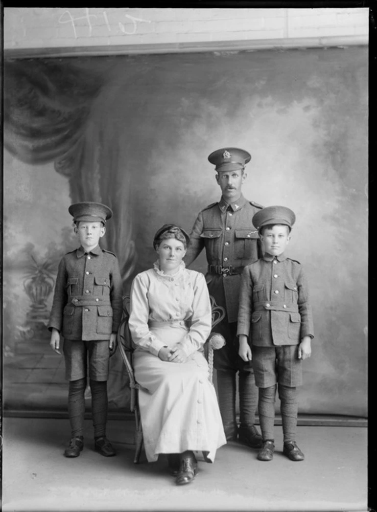 Image: Studio unidentified family portrait, a World War I soldier with moustache, collar and hat badges, standing with two sons in military style uniforms and hats alongside their mother sitting, Christchurch