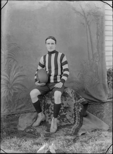Image: Portrait of unidentified male outdoors, in a rugby uniform, holding a ball, with a painted studio backdrop, probably Christchurch district