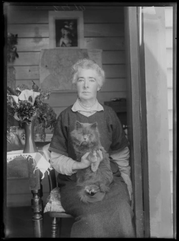 Image: Portrait of an unidentified woman sitting within the doorway of her wooden house holding her cat with a vase of flowers on a table, probably Christchurch region