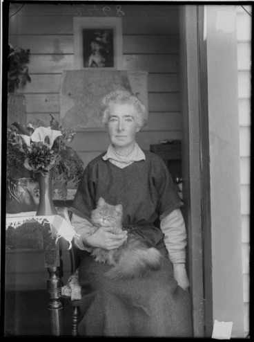 Image: Portrait of an unidentified woman sitting within the doorway of her wooden house with her cat with a vase of flowers on a table, probably Christchurch region
