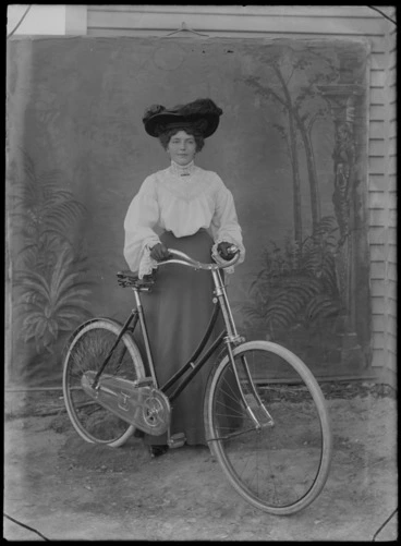 Image: Outdoors in front of a false backdrop, portrait of an unidentified young woman in a high lace v-neck blouse with bar brooch and long dark skirt, standing with a large feathered hat and gloves holding a woman's bicycle, probably Christchurch region
