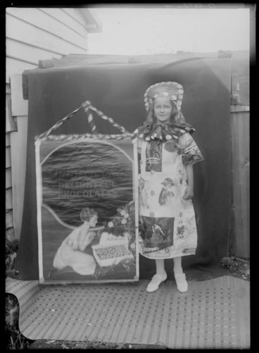 Image: Outdoors by the side of a wooden building, an unidentified young girl in a costume made up with Aulsebrook's Chocolates advertising pictures, standing beside a large advertising sign with 'Aulsebrook's Delicious Chocolates', probably Christchurch