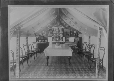 Image: Inside a large tent setup as a dining room, with carpet, a large table with potted plant, chairs, piano, china cabinet with china and sideboard, postcards and framed pictures, [Sumner?], Christchurch